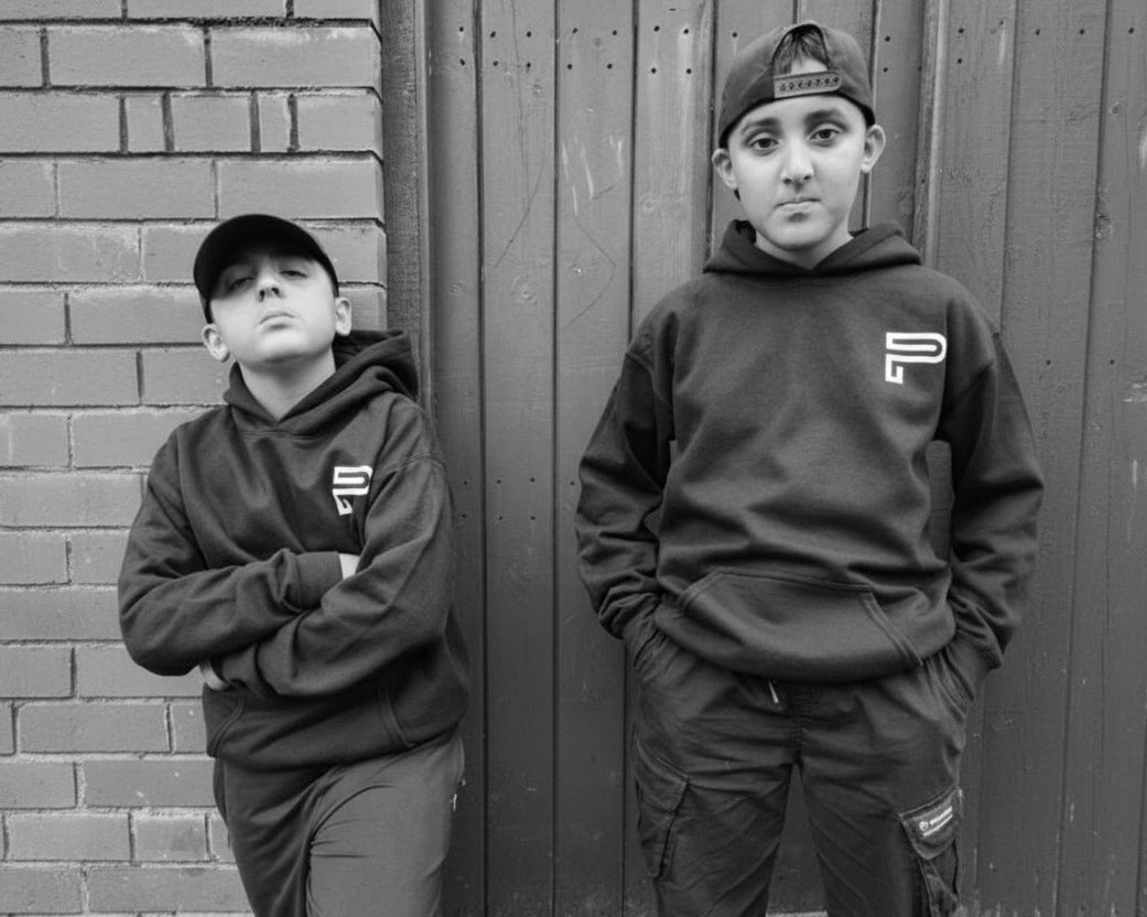 Two children wearing matching black hoodies with a logo against a brick wall.